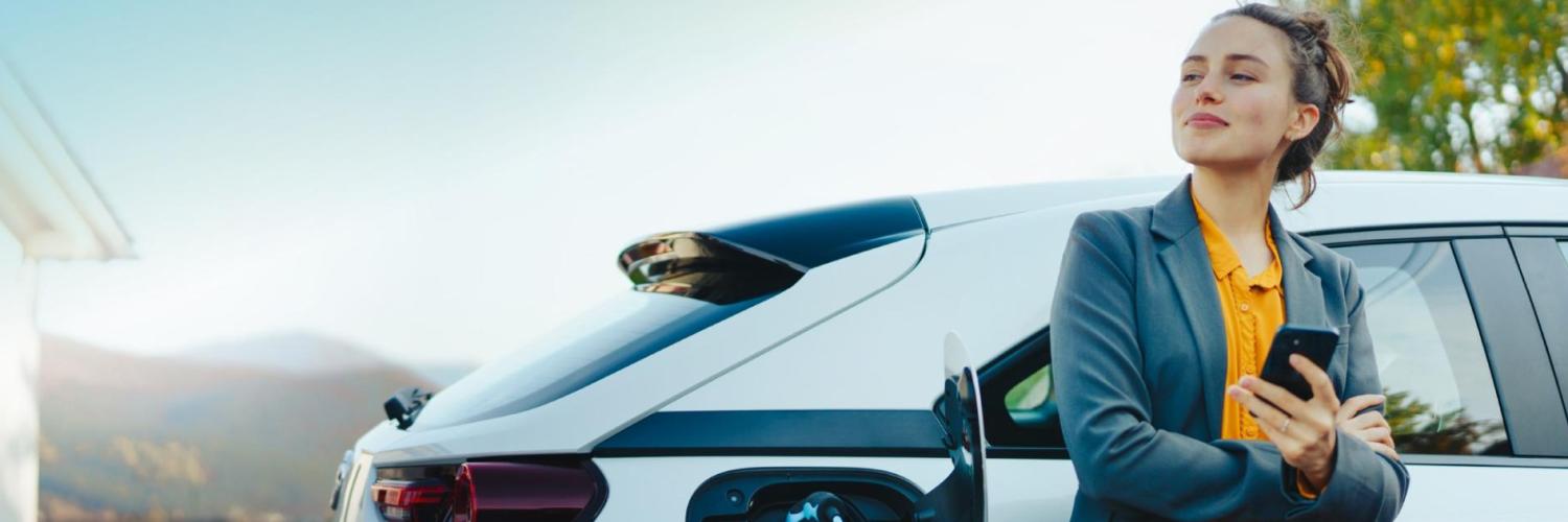 Smilende dame som lener seg på en elbil koblet til lader. 